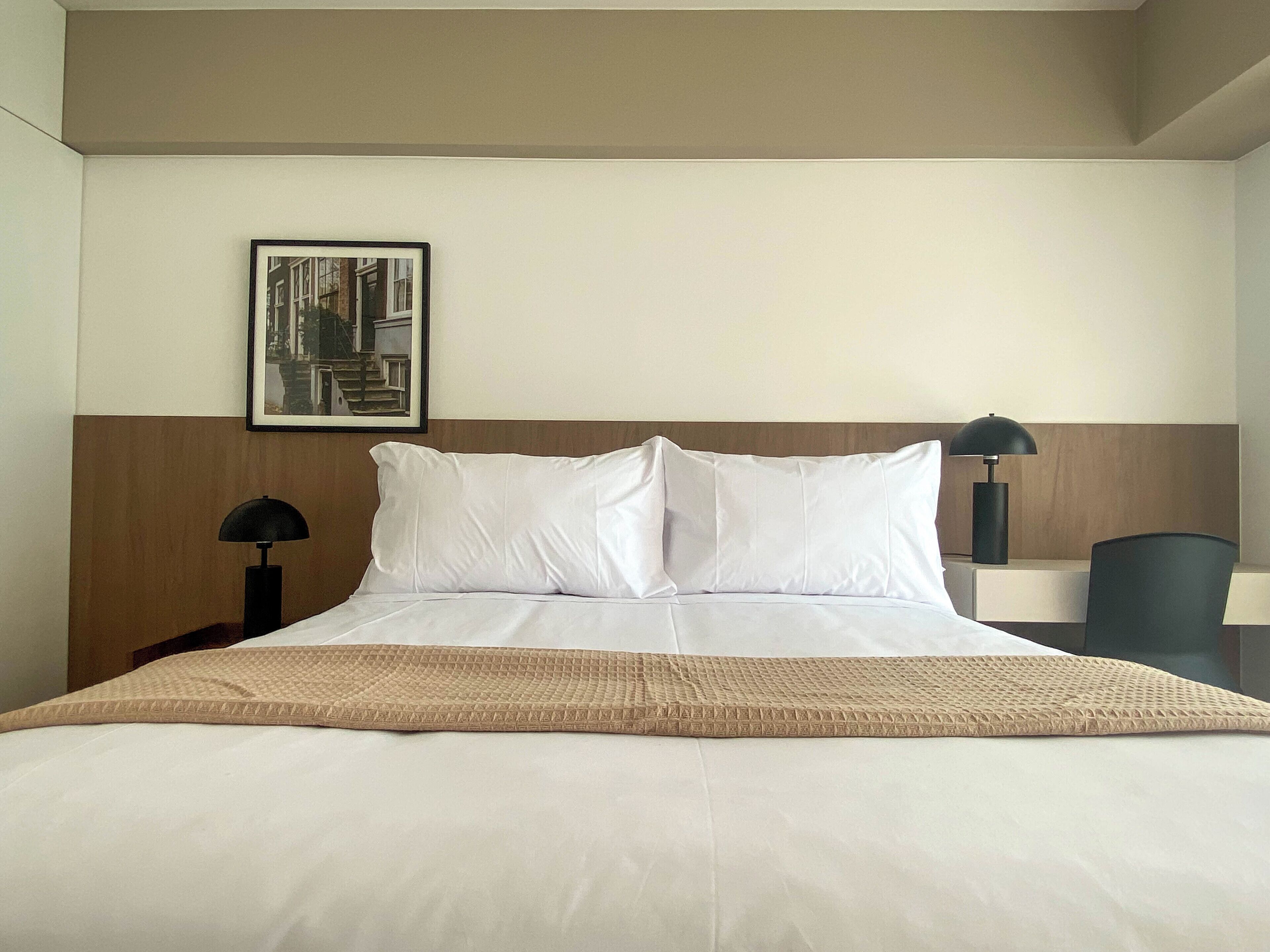 Una habitación de hotel con una cama, dos mesitas de noche, una lámpara, una silla y un cuadro en la pared.