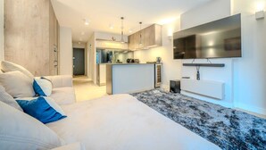 A modern living room with a sofa, a flat-screen TV mounted on the wall, and a kitchen area in the background. - The Nothinghill Charmer (London)