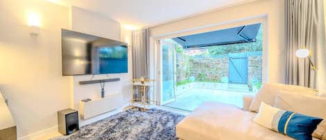 A modern living room with a flat-screen TV, a sofa, a coffee table, and a large window offering a view of a garden.