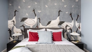 A bedroom with a bed, red and grey pillows, a nightstand with a lamp, and a wall with a bird mural. - Lucky Cranes (Bucharest)
