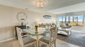 Un coin repas avec une table et des chaises en verre, un lustre, un miroir décoratif et, au fond, une enseigne sur laquelle on peut lire « SABLE ET VAGUES ».