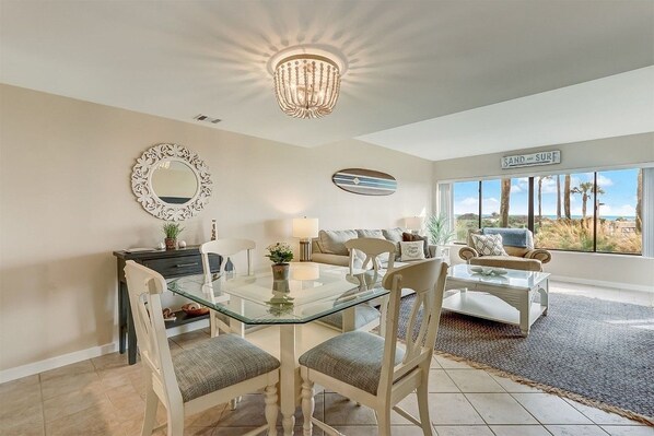 A dining area with a glass table and chairs, a chandelier, a decorative mirror, and a sign reading "SAND AND SURF" in the background.