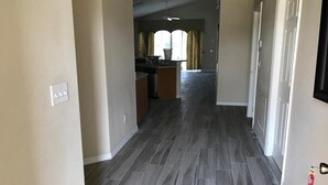 Un couloir avec un coin cuisine au fond, doté d’un parquet en bois foncé et d’une fenêtre avec des rideaux.