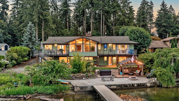 A two-story house with a dock extending into a calm body of water, surrounded by dense forest. - Lakefront Home with Hot Tub, Kayaks, Fire Pit, & Breakfast Included (Black Diamond)