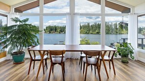 A dining area with a large table and chairs, large windows with curtains, and potted plants. - Lakefront Home with Hot Tub, Kayaks, Fire Pit, & Breakfast Included (Black Diamond)