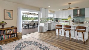 A modern kitchen with white cabinets, a central island with bar stools, a dining area with a table and chairs, and a bookshelf with books. - Lakefront Home with Hot Tub, Kayaks, Fire Pit, & Breakfast Included (Black Diamond)