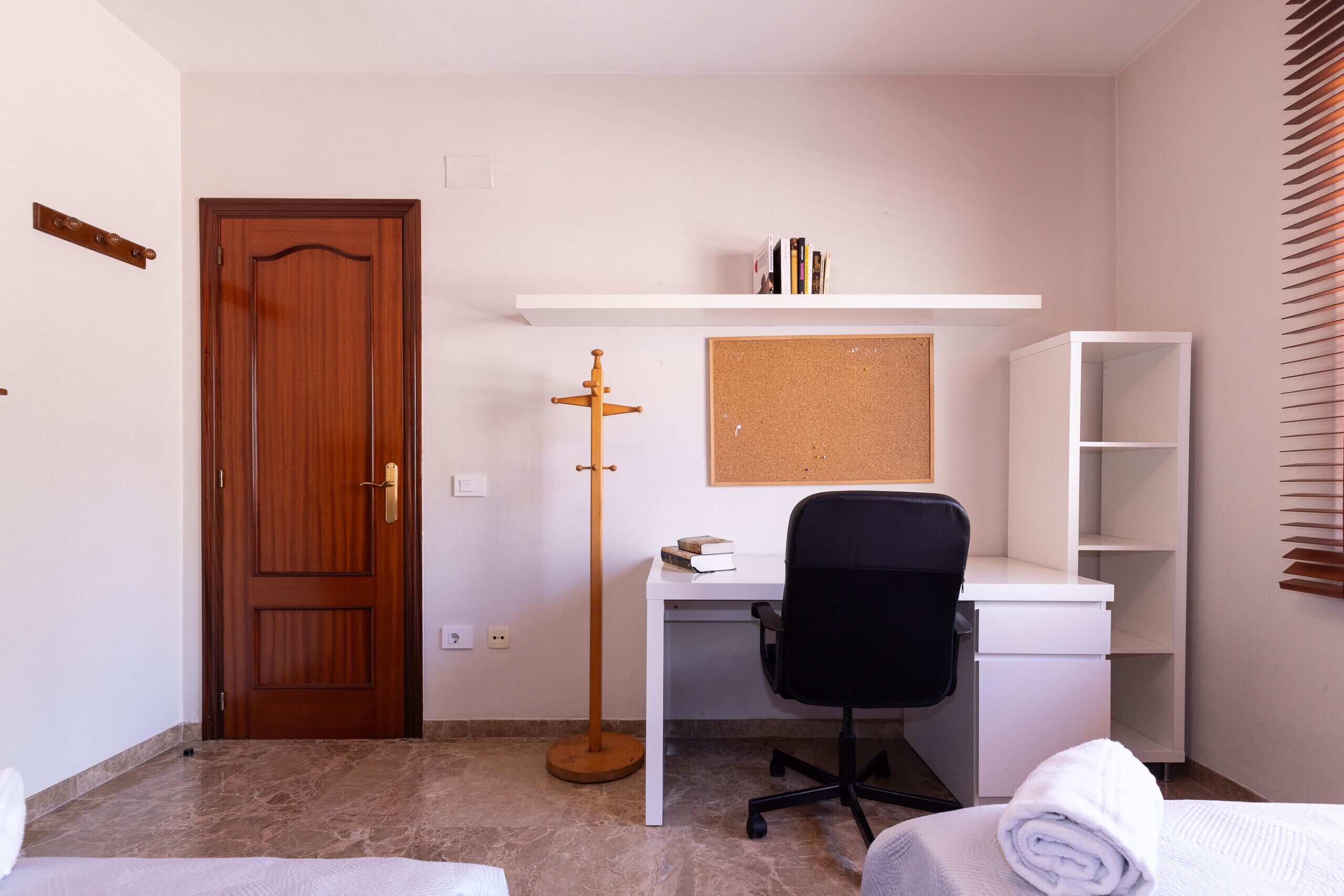 Una habitación con puerta de madera, un escritorio blanco con una silla de oficina negra, una pizarra de corcho y un estante blanco con libros.