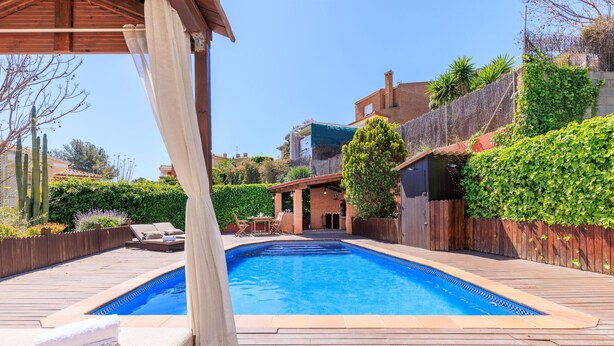 Una piscina con bordo in legno, una zona relax coperta e vista su un quartiere residenziale con un edificio in mattoni e vegetazione.