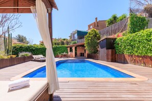 A swimming pool with a wooden deck, a covered seating area, and a view of a residential area with a brick building and greenery. - Large Villa between the beach and the forest (Tarragona)
