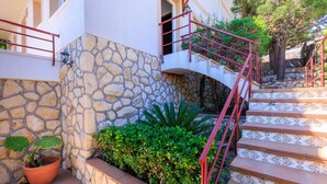 Una scala in pietra con ringhiere rosse che conduce all'ingresso di un edificio, circondata da piante in vaso e vegetazione.