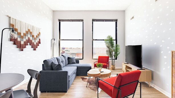 A modern living room with a grey sectional sofa, a red chair, a round wooden coffee table, a TV stand, and a large window with a city view. - Cleveland Loft-Wolstein/Progressive/Playhouse/CSU. (Cleveland)