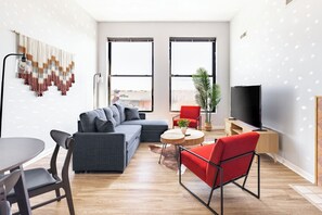A modern living room with a grey sectional sofa, a red chair, a round wooden coffee table, a TV stand, and a large window with a city view. - Cleveland Loft-Wolstein/Progressive/Playhouse/CSU. (Cleveland)
