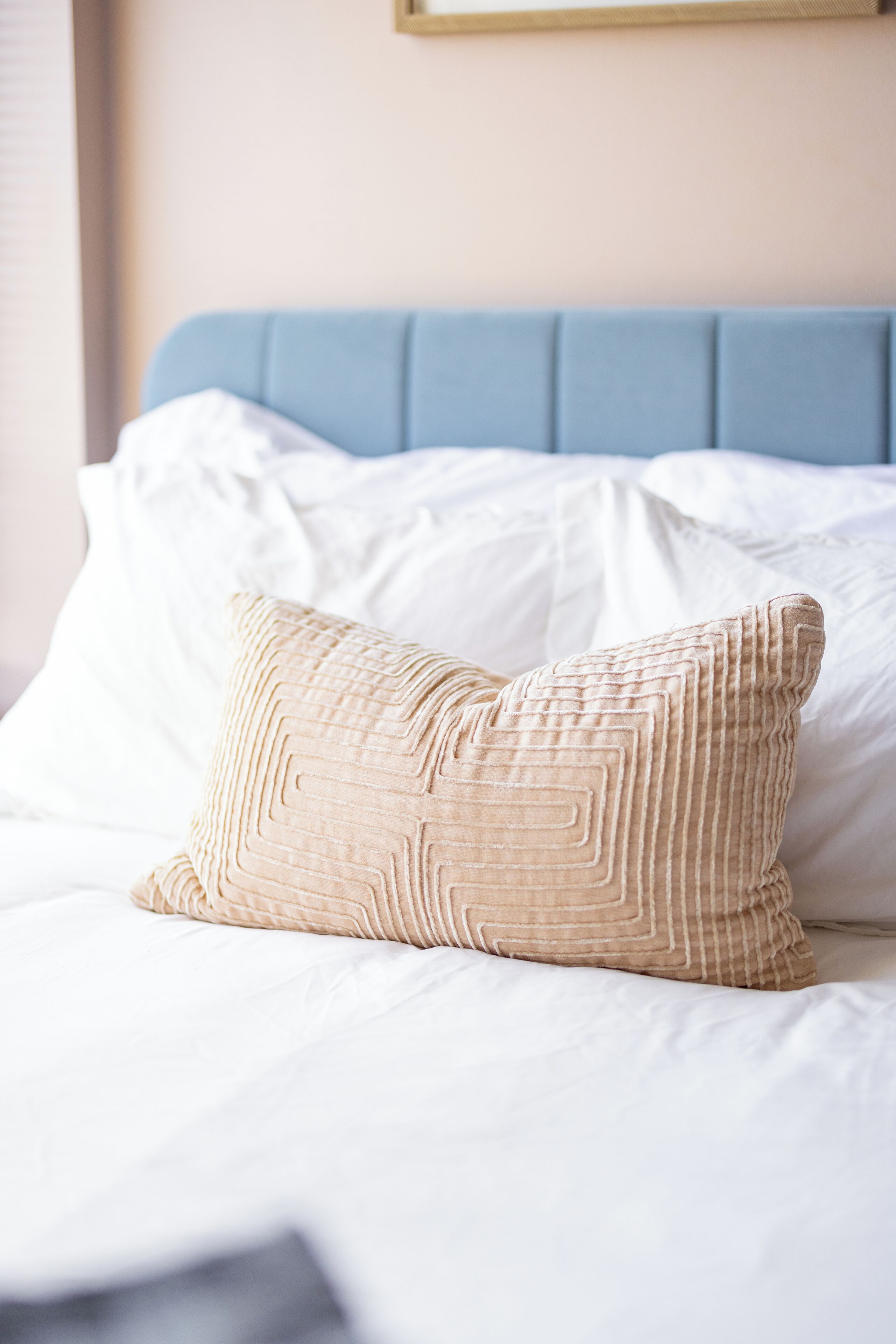 Uma cama com cabeceira azul, roupa de cama branca e almofada bege estampada com um padrão geométrico.