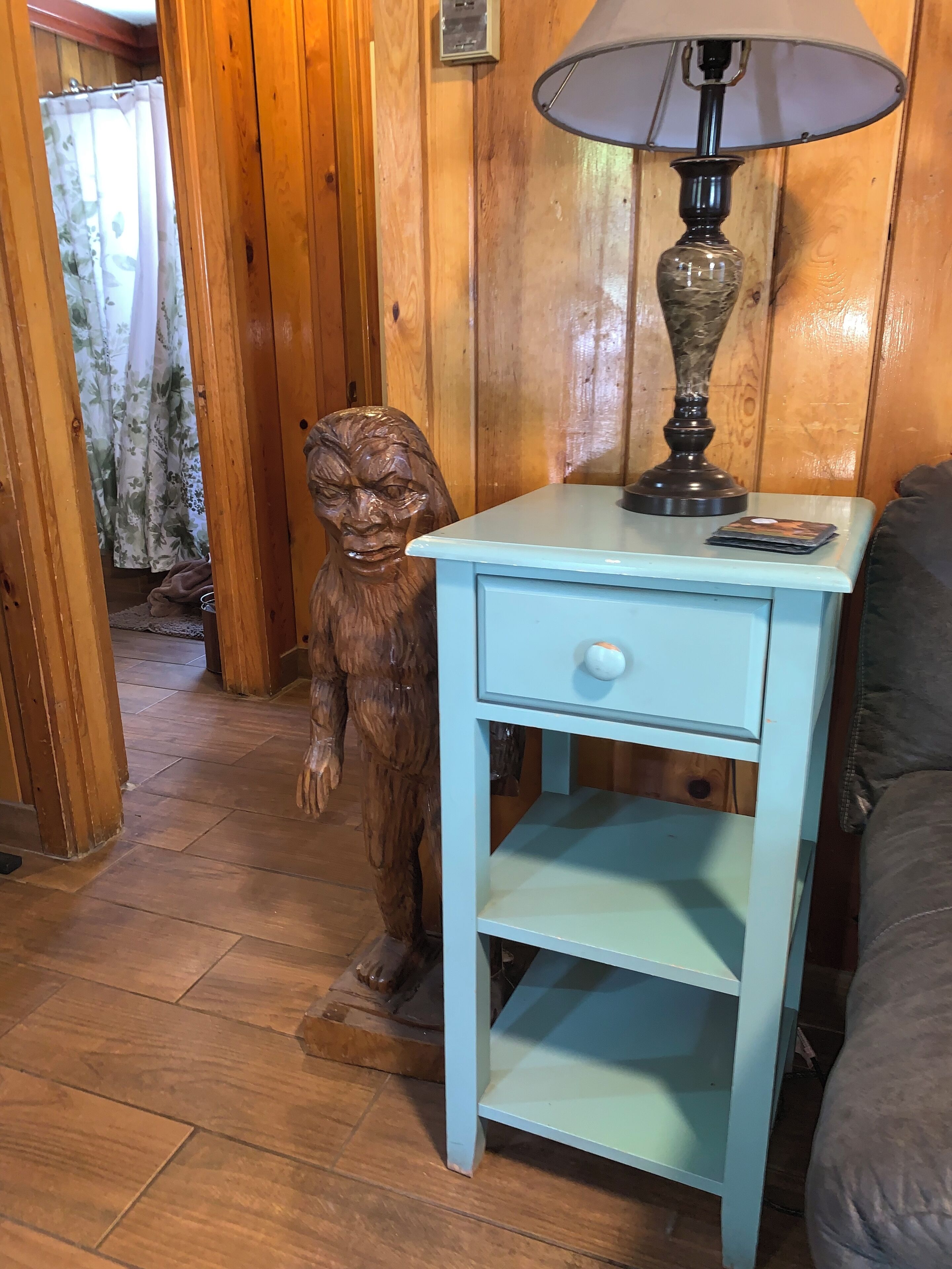 Une pièce aux murs lambrissés, avec une table de chevet bleue, une lampe et une statue en bois.