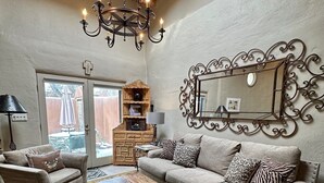 A cozy living room with a wooden ceiling, a chandelier, a sofa set, a coffee table, and a large mirror with ornate metalwork. - Elegant casita in the middle of Santa Fe's historic Canyon Road district  (Santa Fe)