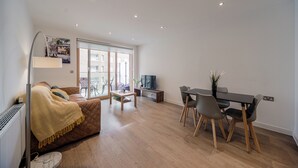 A modern living room with a sofa, a dining table with chairs, a TV, and a large window with a view of buildings. - Barking Bright Apartment With 2 Bedrooms (Barking)