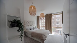 A modern bedroom with a large bed, a white headboard, a plant on a shelf, and a window with curtains. - Barking Bright Apartment With 2 Bedrooms (Barking)