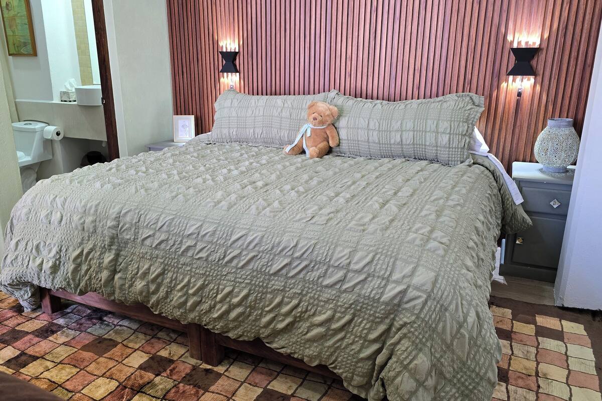 Una cama bien hecha con una colcha acolchada, un oso de peluche sobre las almohadas y una mesita de noche con un jarrón decorativo.