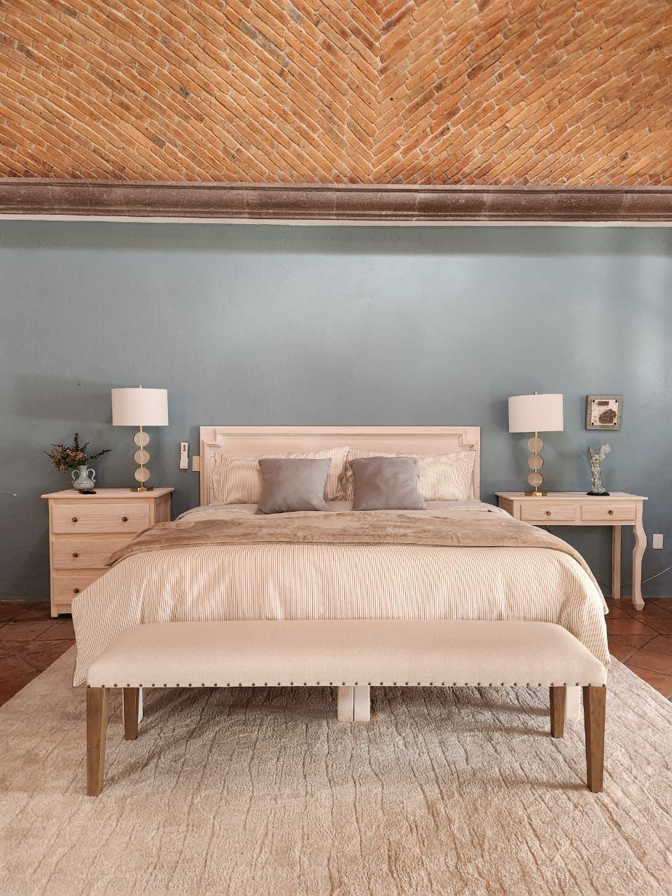 A bedroom with a herringbone-patterned ceiling, a bed with a bench, two bedside tables with lamps, and a wall-mounted picture frame.