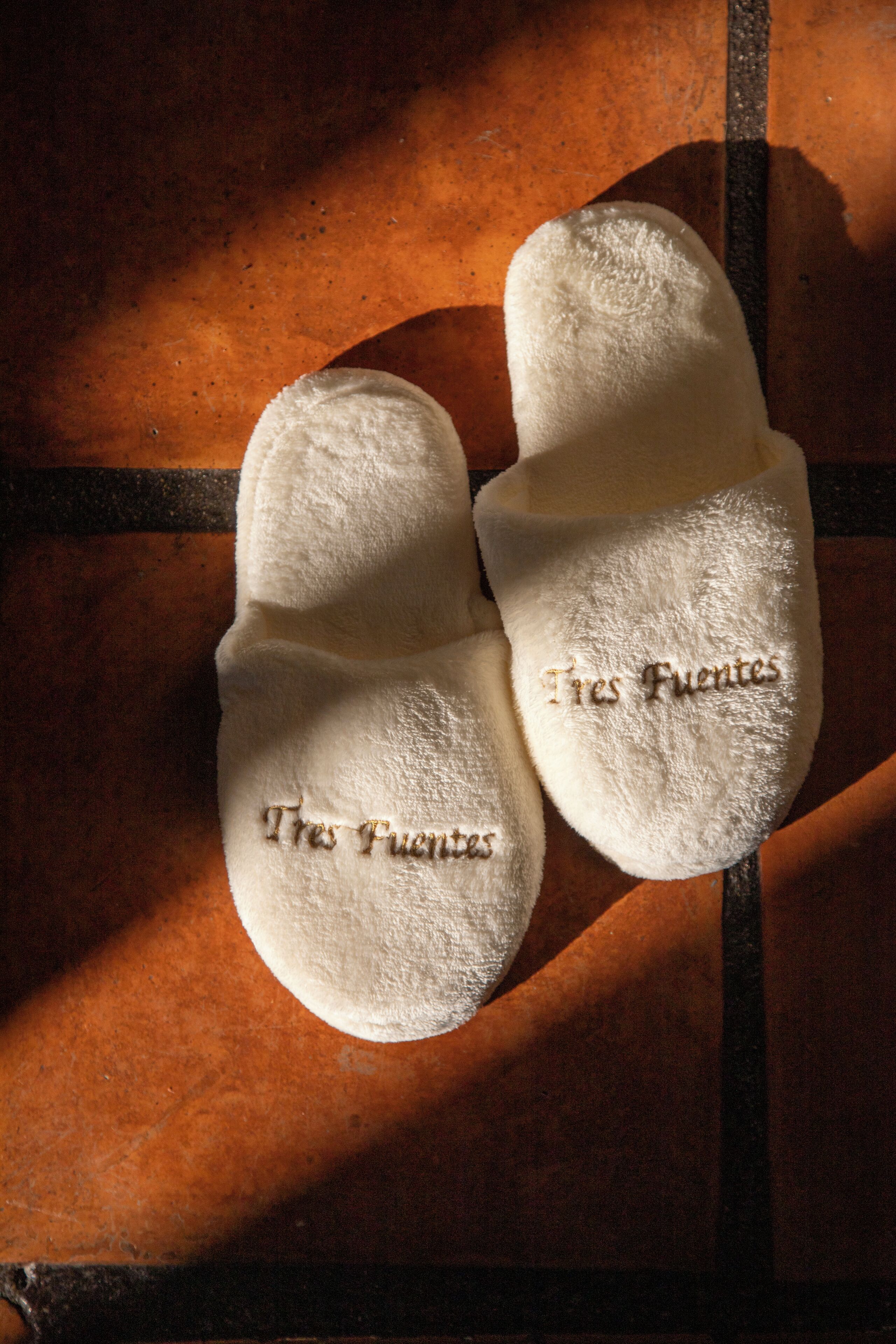 A pair of white slippers with embroidered text "Tres Fuentes" on a textured surface.