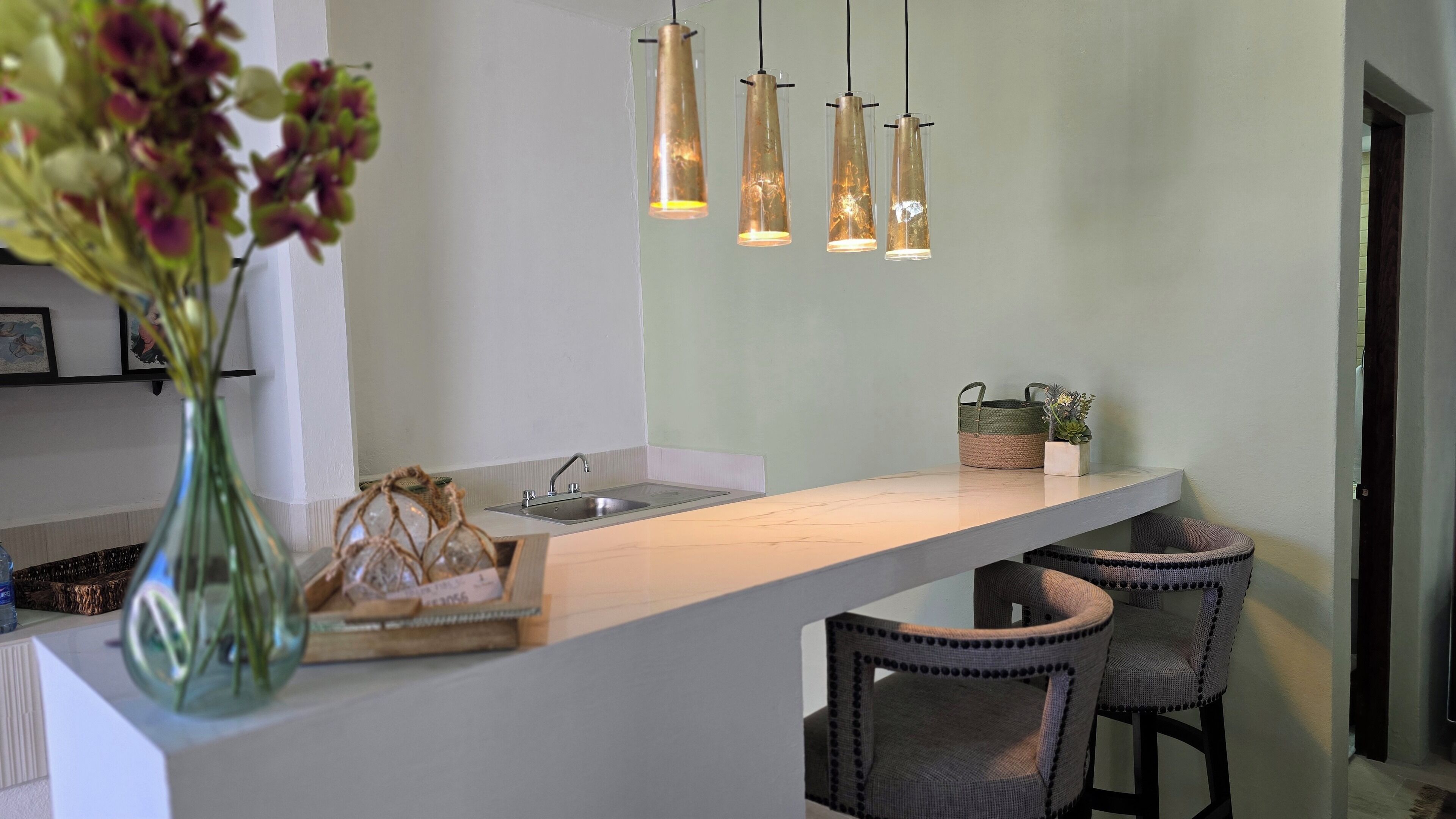 A modern kitchen with a white countertop, a sink, and hanging pendant lights. There are two wicker chairs and a small potted plant on the counter.