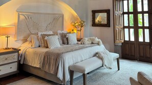 Un dormitorio con techo de ladrillo, una cama grande con varios almohadones, una mesita de noche, un banco y una lámpara colgante.