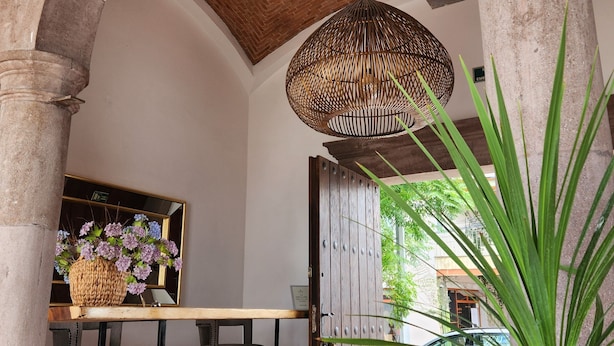 Un interior con un arco de piedra, una lámpara de mimbre colgante, una mesa de madera con sillas y un espejo grande.