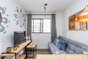 A modern living room with a flat-screen TV, a wooden table, a sofa with cushions, a large window with curtains, and a pendant light. - Edifício Lins 236 (Curitiba)