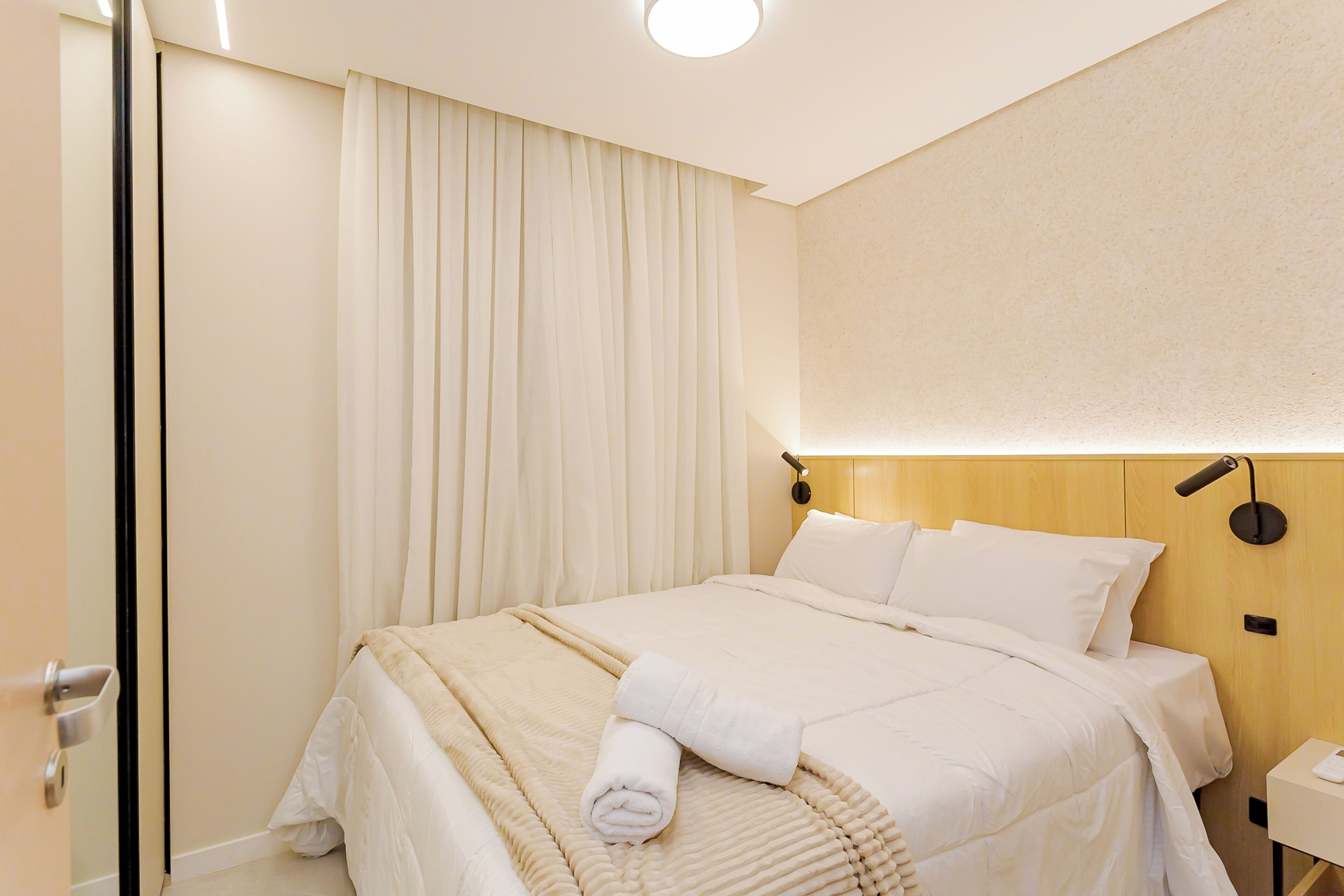 A neatly made bed with white linens, a bedside table with a lamp, and a curtain covering a window.