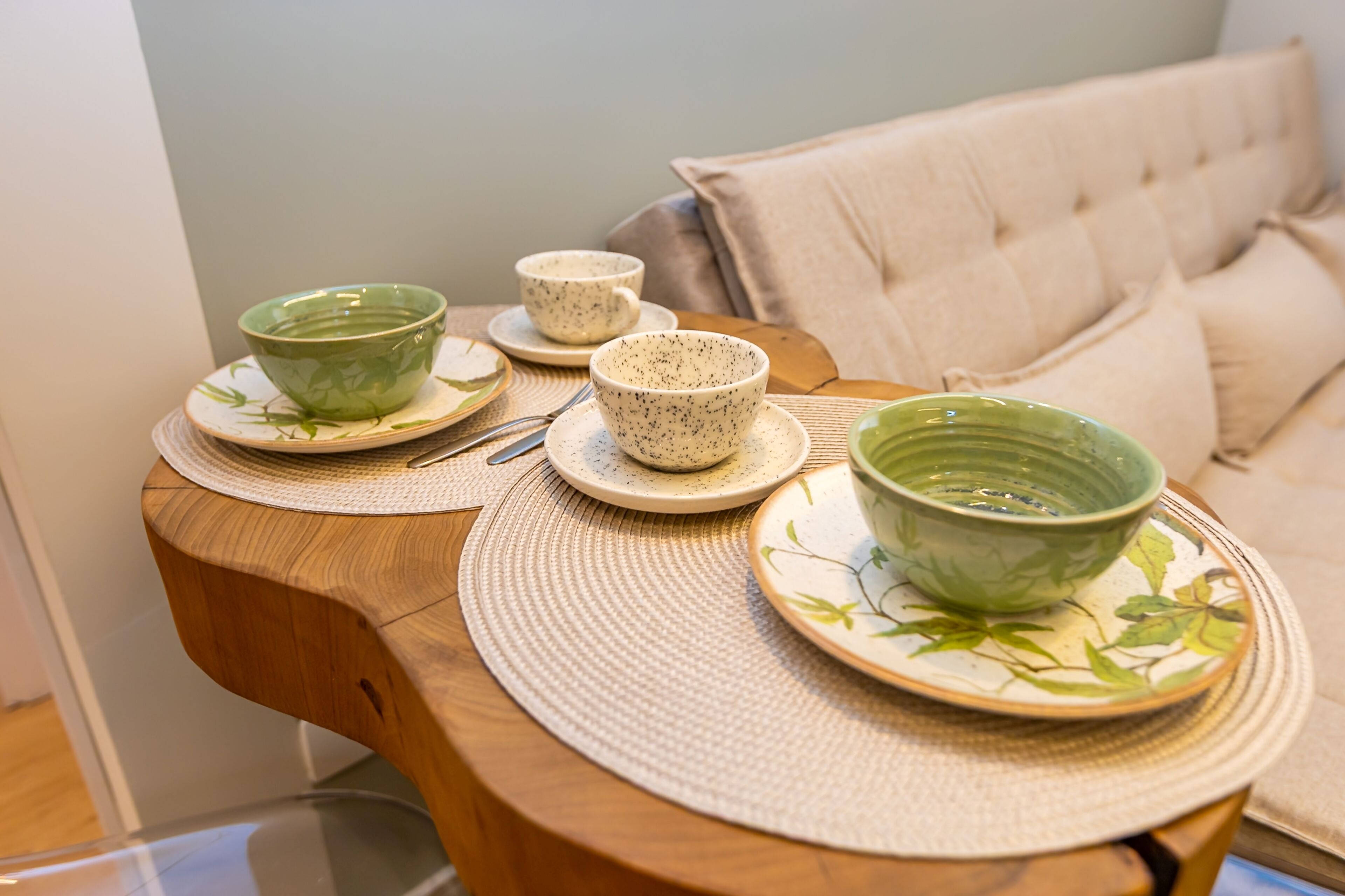 Une table en bois, sur laquelle sont posés des bols et des assiettes en céramique, un set de table en fibres tressées, et un canapé en arrière-plan.