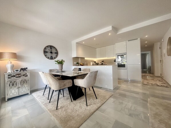 A modern kitchen with a central island, a dining area with a round table and chairs, and a living space with a sofa and a lamp. - K13 Ocean Point - Apartment (Sitio de Calahonda)
