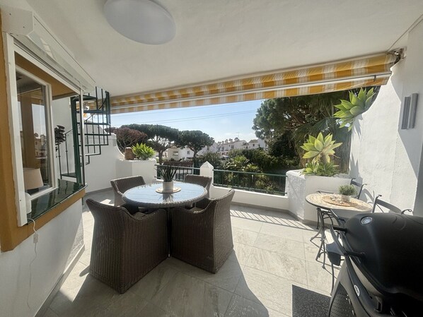 A balcony with a round table and wicker chairs, a grill, and potted plants.