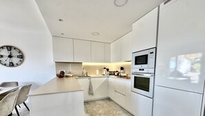 A modern kitchen with white cabinetry, a central island, a microwave, and a clock on the wall. - K13 Ocean Point - Apartment (Sitio de Calahonda)