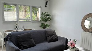 Ein modernes Wohnzimmer mit einem dunkelgrauen Sofa, einem weißen Couchtisch und einem kleinen weißen Tisch mit einer Blumenvase. Es gibt ein Fenster mit Jalousien, eine Pflanze und eine weiße Kommode mit Spiegel.