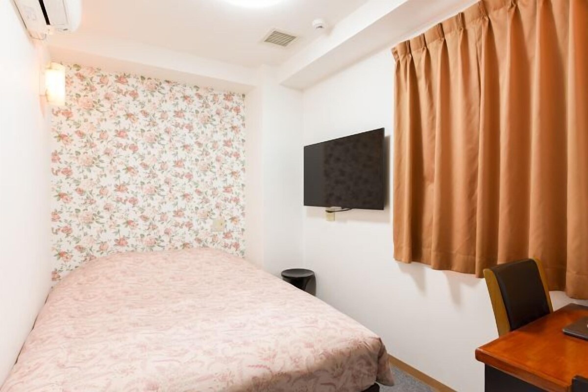 A bedroom with a bed, a desk, a chair, a TV mounted on the wall, and a window with curtains.