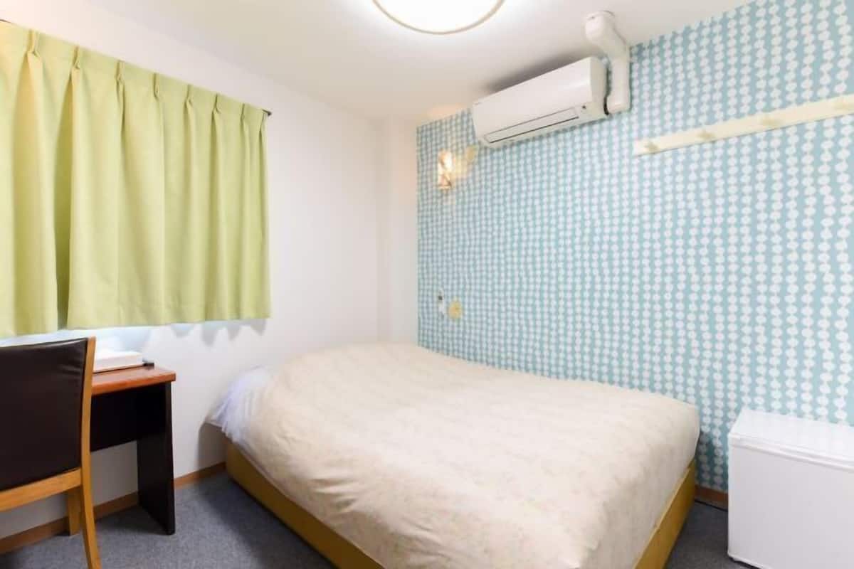 A small, neatly arranged room with a bed, a desk, and a chair. An air conditioning unit is mounted on the wall above the bed.