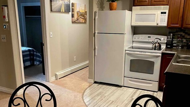 A compact kitchen with a refrigerator, stove, microwave, and wooden cabinets. A dining table with chairs is in the foreground.