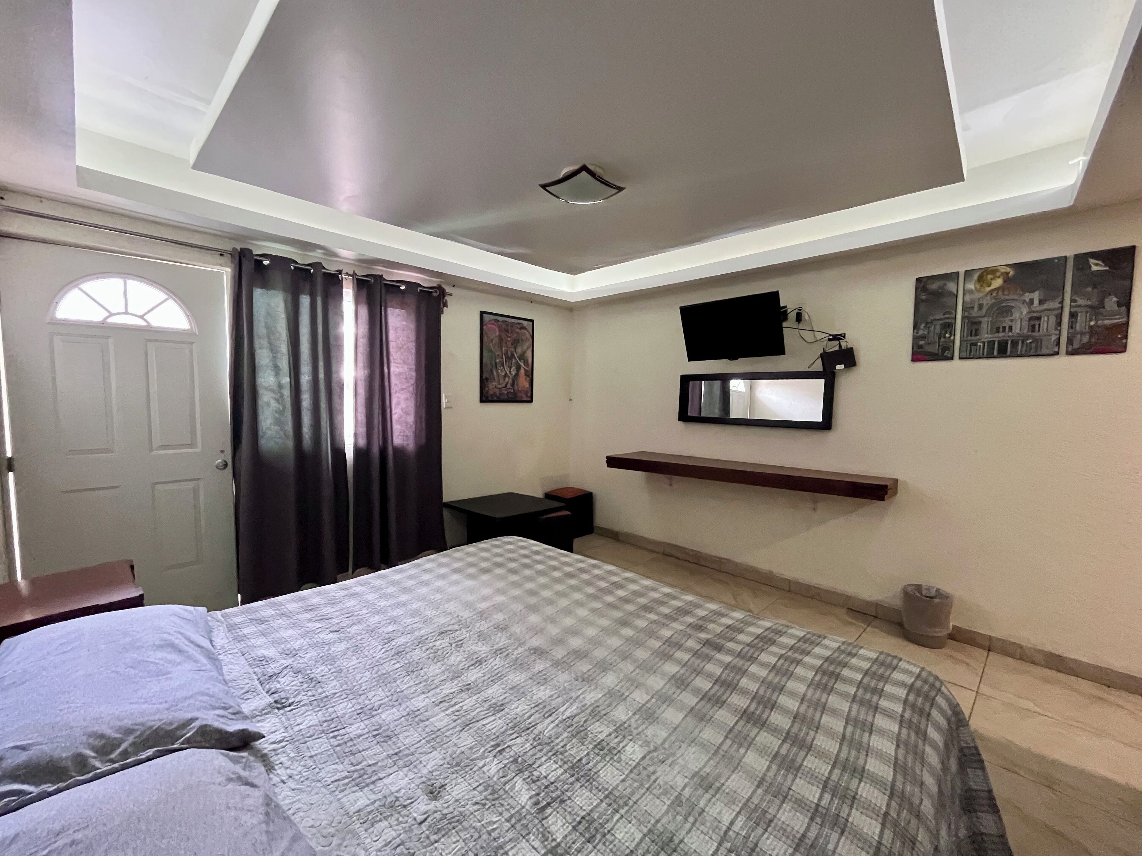 A bedroom with a bed, a desk, a TV mounted on the wall, and a window with curtains.