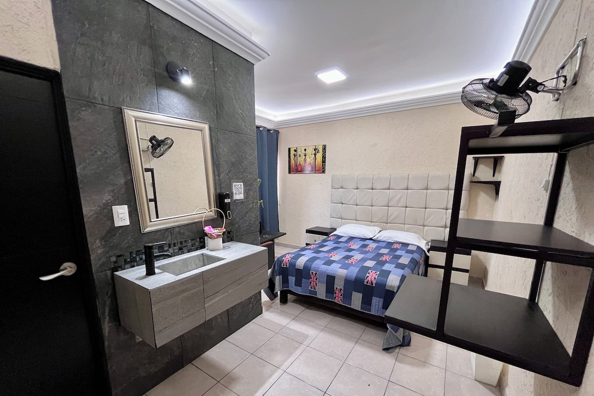 A modern bedroom with a large bed, a bedside table, a nightstand, a built-in shelf, a mirror, and a bathroom vanity with a sink and towel rack.