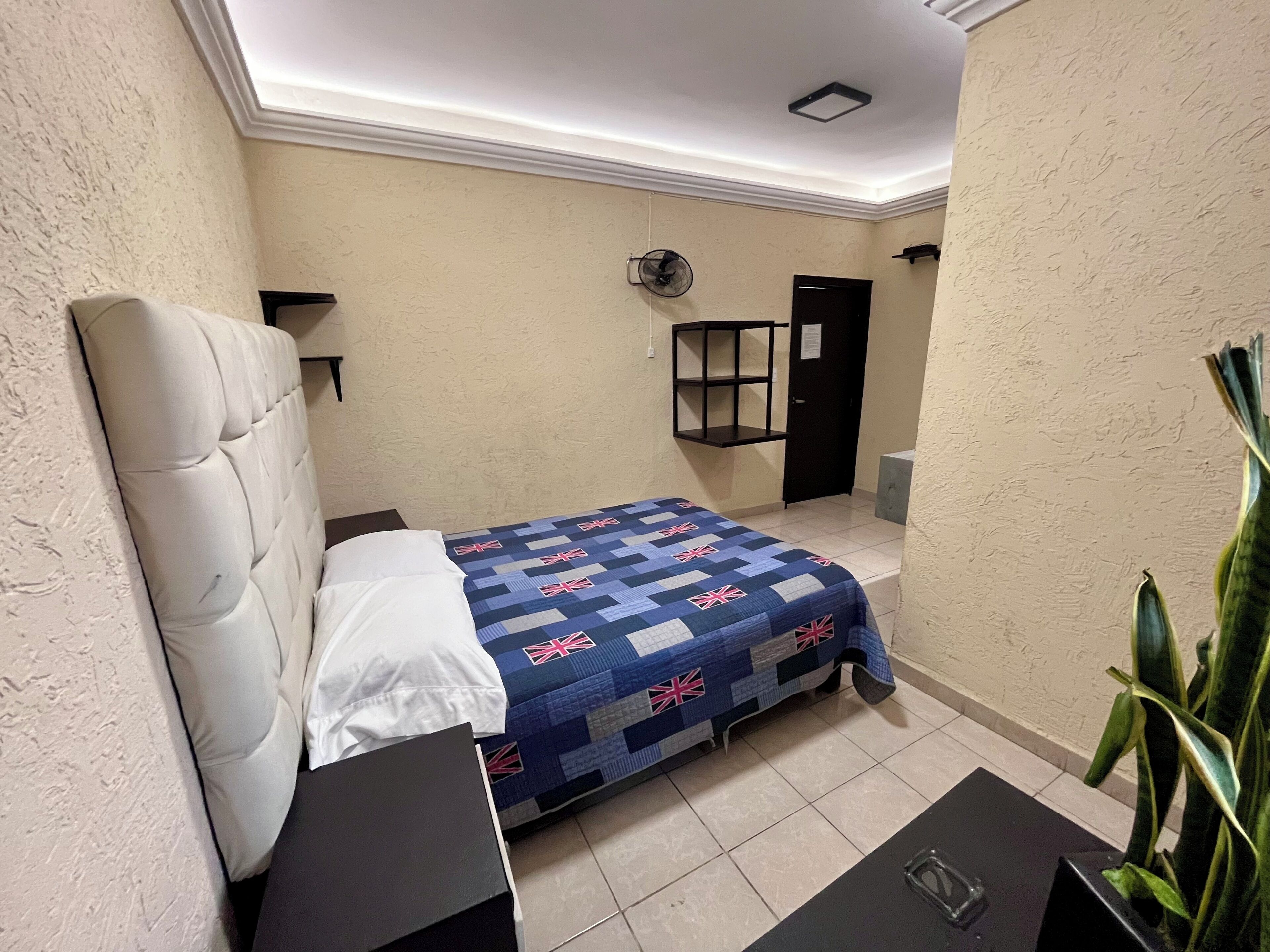 Un dormitorio con cama, mesita de noche, estante, puerta y ventilador.