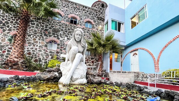 A statue of a woman sitting by a pond with lily pads, surrounded by a stone building with arched windows and a blue building with a door marked "10". - Hotel El Castillo (Cuernavaca)