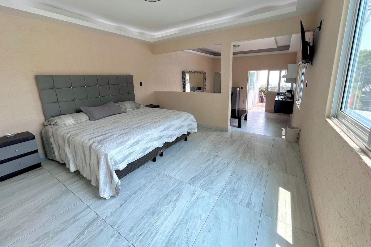 A bedroom with a large bed, a nightstand, a dresser, a mirror, and a view of the outside.