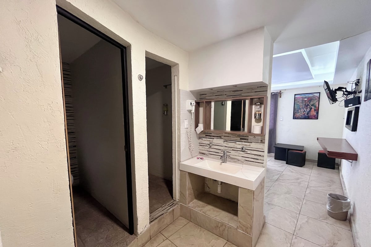 A modern bathroom with a large mirror, a sink, and a shower. There is a door leading to another room, and a small trash bin is visible.