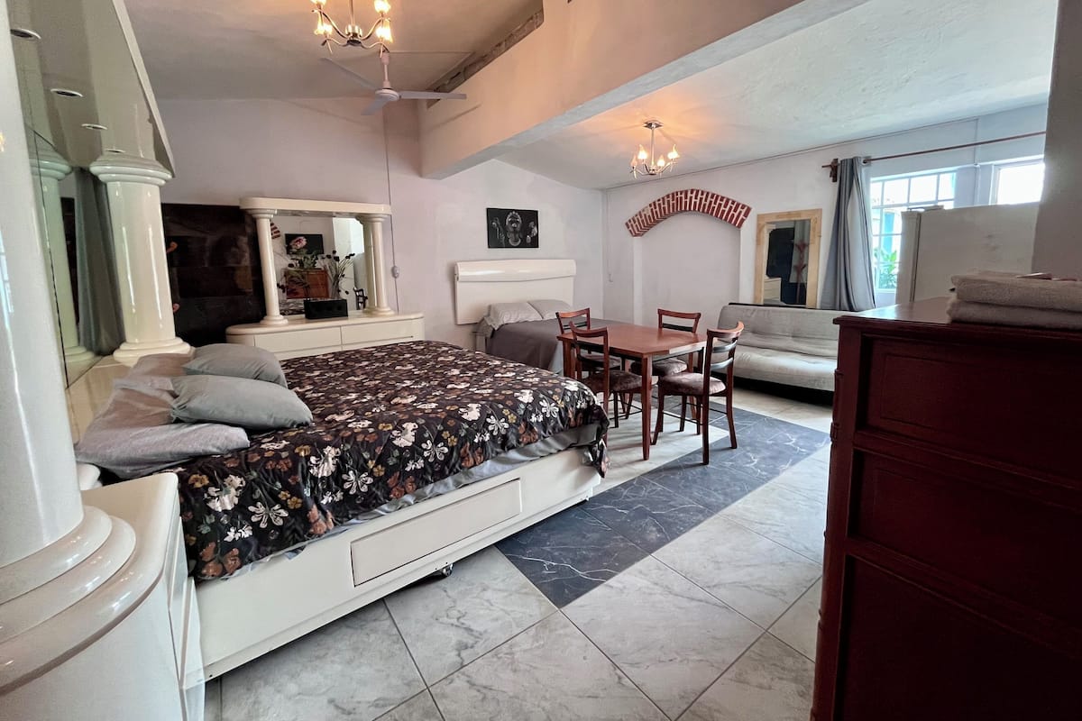 A bedroom with a bed, bedside tables, a dresser, a dining table with chairs, a sofa, and a chandelier.