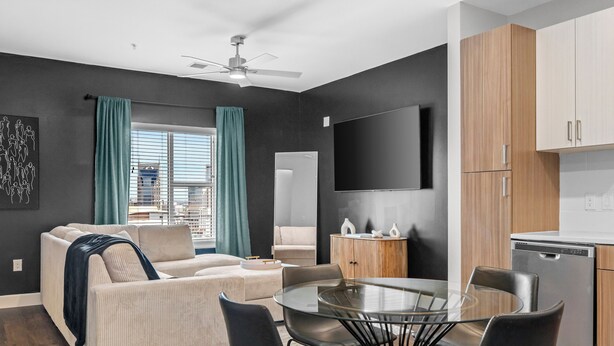 Ein modernes Wohnzimmer mit einer Couch, einem Glastisch und schwarzen Stühlen. Es gibt eine Küchenzeile mit Holzschränken und einem Geschirrspüler.