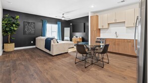 Ein modernes Wohnzimmer mit Sofa, Esstisch und Stühlen. Es gibt eine Küchenzeile mit Holzschränken und einem Kühlschrank. Eine große Pflanze steht in einem gewebten Korb, und eine Deckenventilator ist zu sehen.