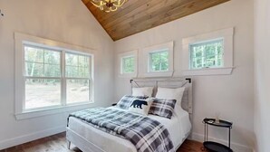 A bedroom with a large bed, multiple windows, a wooden ceiling, and a small side table. - NEW BUILD! Mountain views, minutes from Squam's Sandwich Beach and boat ramp!  (Center Sandwich)