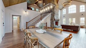 A spacious dining area with a long wooden table, surrounded by wooden chairs, a stone fireplace, and a staircase leading to an upper level. - NEW BUILD! Mountain views, minutes from Squam's Sandwich Beach and boat ramp!  (Center Sandwich)