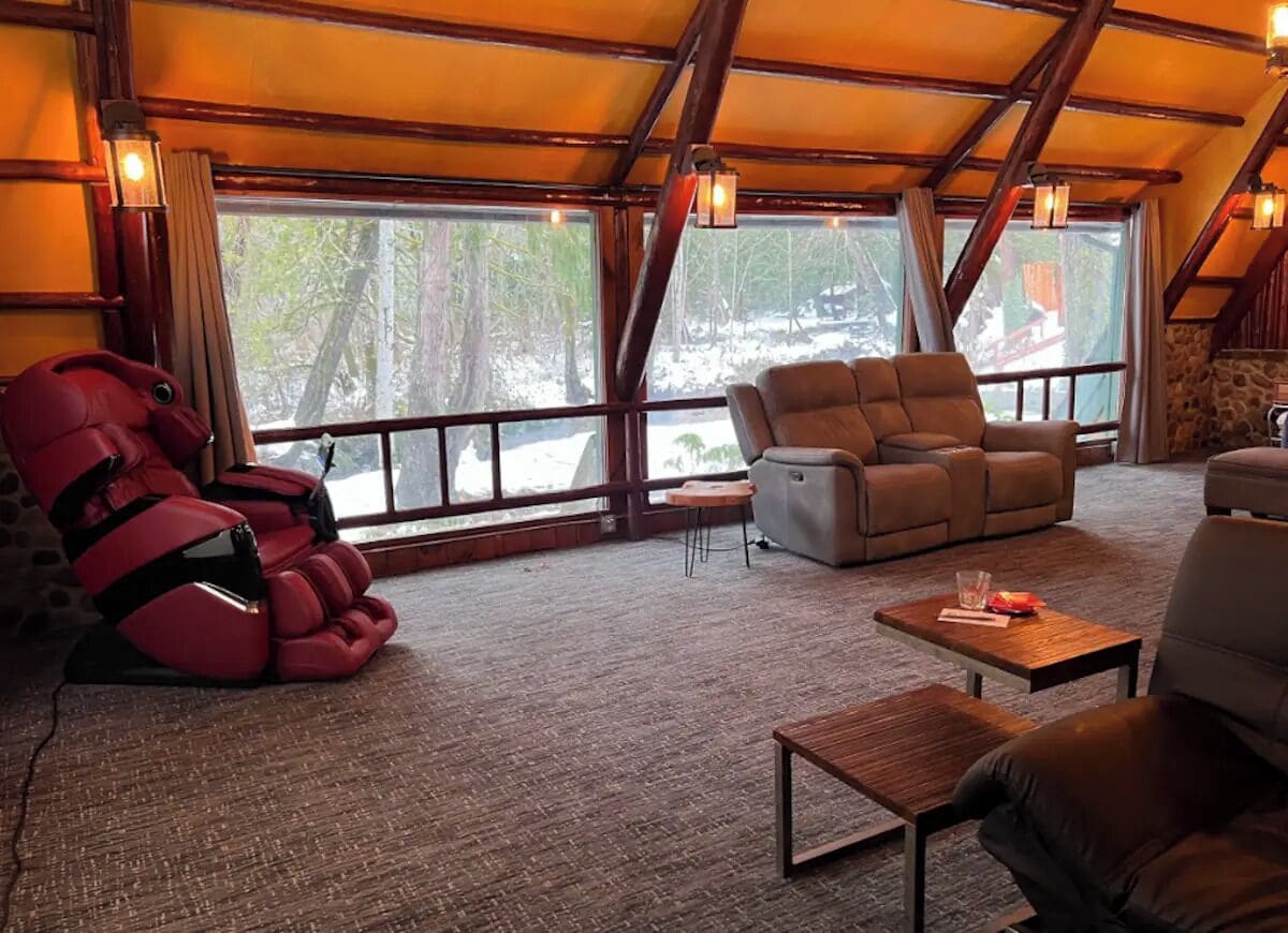 Una habitación acogedora con sillón de masaje, sofá y una mesita. Amplias ventanas ofrecen vistas al exterior.