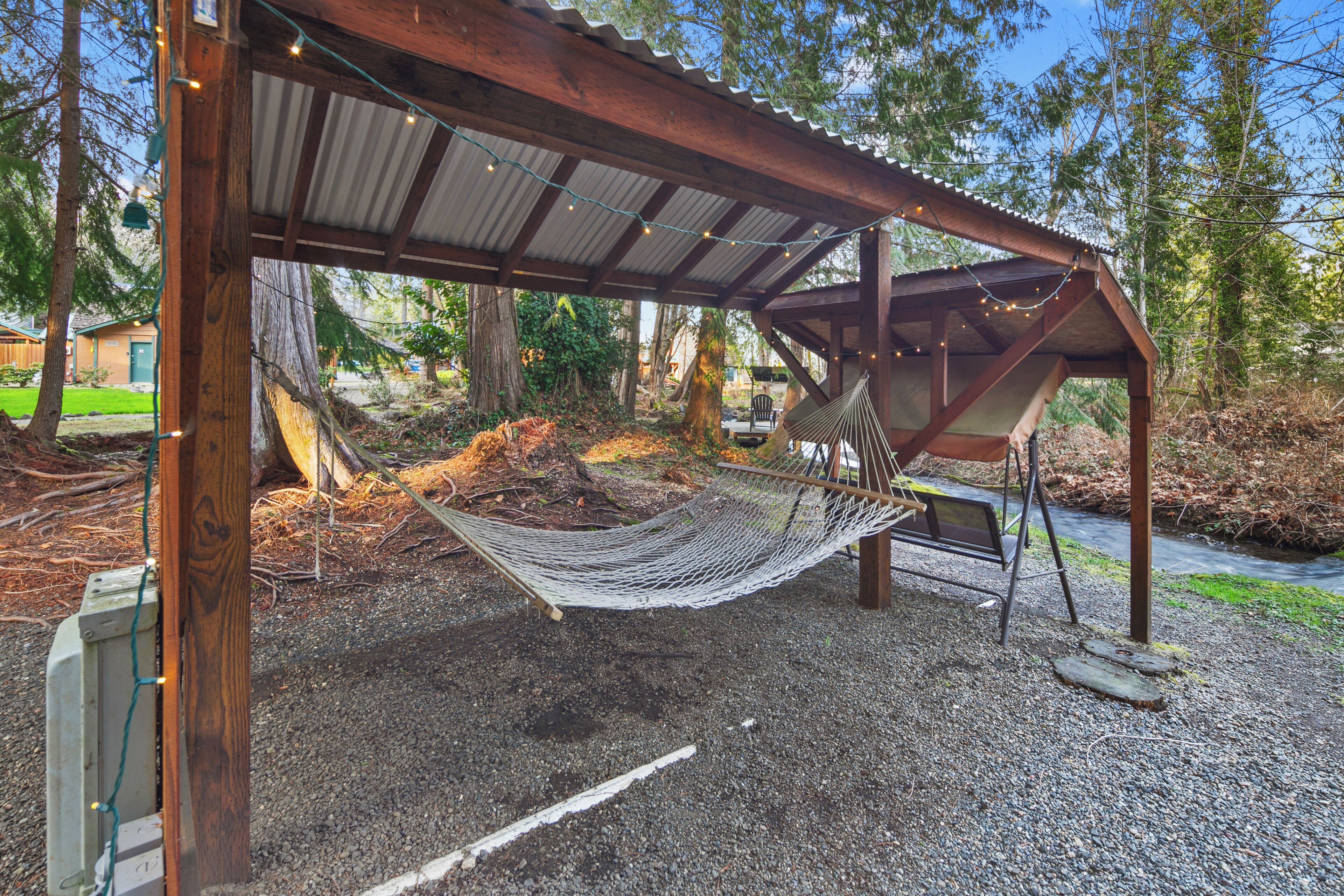 Una hamaca bajo una pérgola de madera con luces colgantes, rodeada de árboles y un sendero de grava.